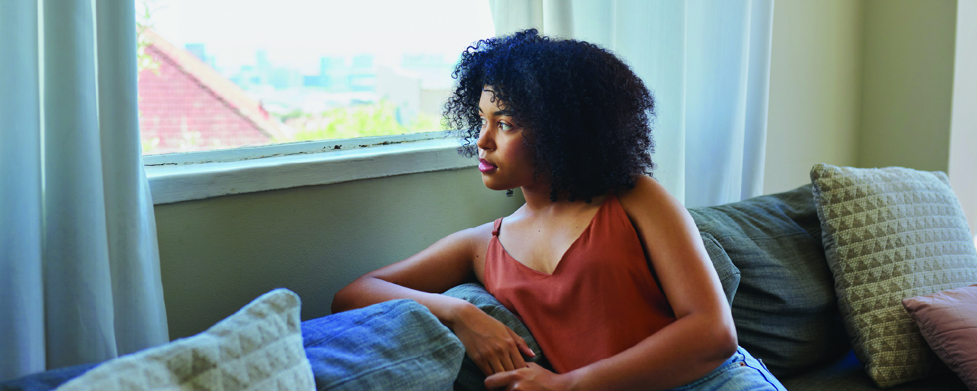 A person with curly hair sits on a couch beside a bright window, gazing thoughtfully outside. Soft natural light fills the living room, creating a quiet, reflective atmosphere with neutral-toned pillows and curtains in the background.