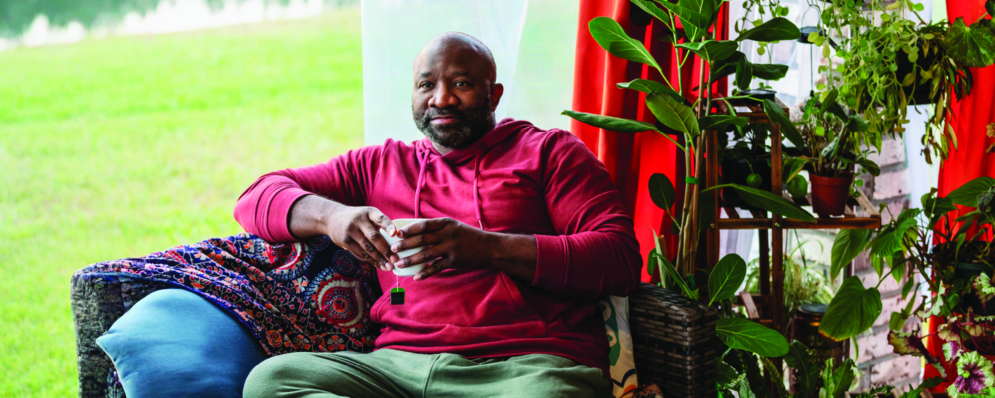 A man in a red hoodie sits comfortably on a sofa holding a mug, surrounded by leafy houseplants beside a bright window.