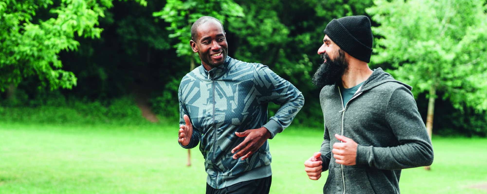 Two adults jogging side by side on a paved path in a green park, smiling and talking as they run. One wears a patterned gray athletic jacket and black leggings, while the other wears a dark beanie, gray hoodie, and blue shorts. Trees and grass fill the background.