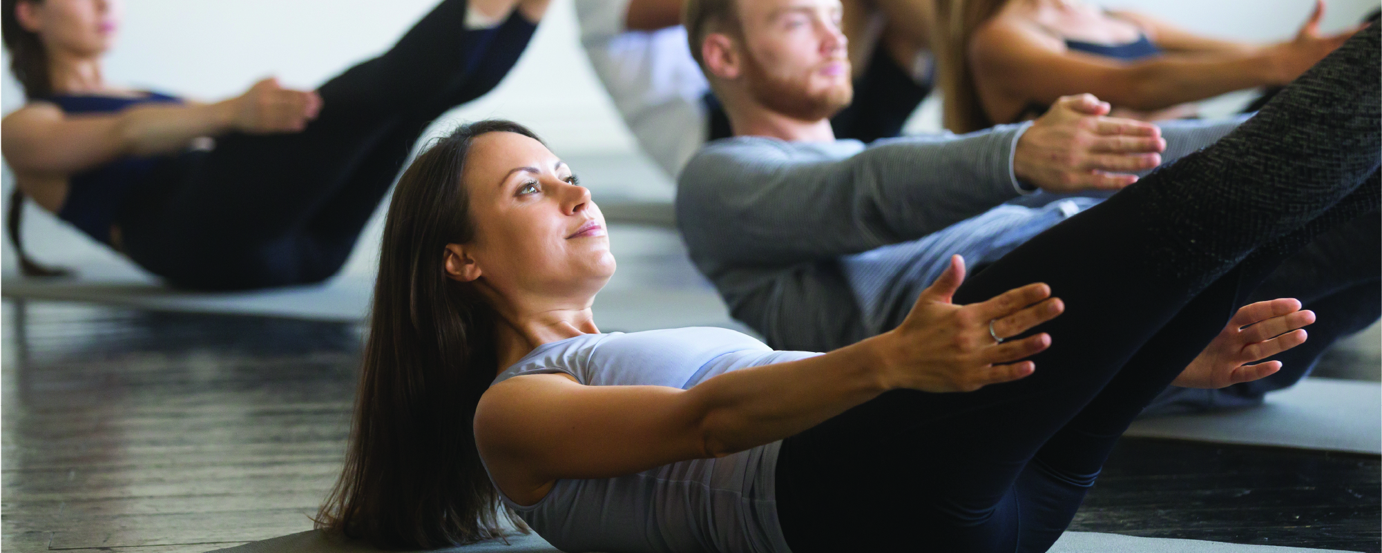A group fitness class performs core-strengthening exercises on yoga mats, with participants holding a V-sit position with arms extended and legs raised in a bright exercise studio.