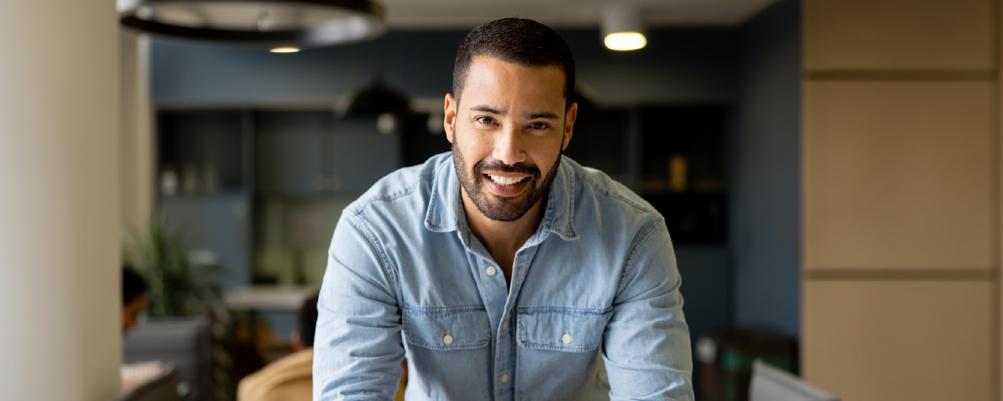 Smiling man in a light blue button-down shirt leans on a kitchen island with folded hands in a modern home, looking directly at the camera.