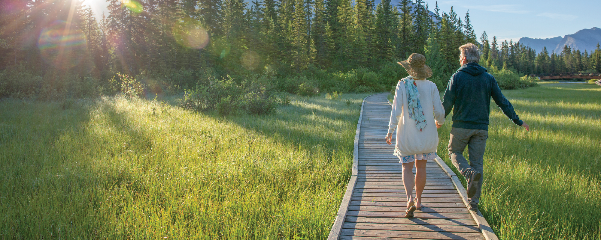 Two people walk hand in hand along a wooden boardwalk through a sunlit mountain meadow, surrounded by evergreen trees with rugged peaks rising in the background under a clear blue sky.