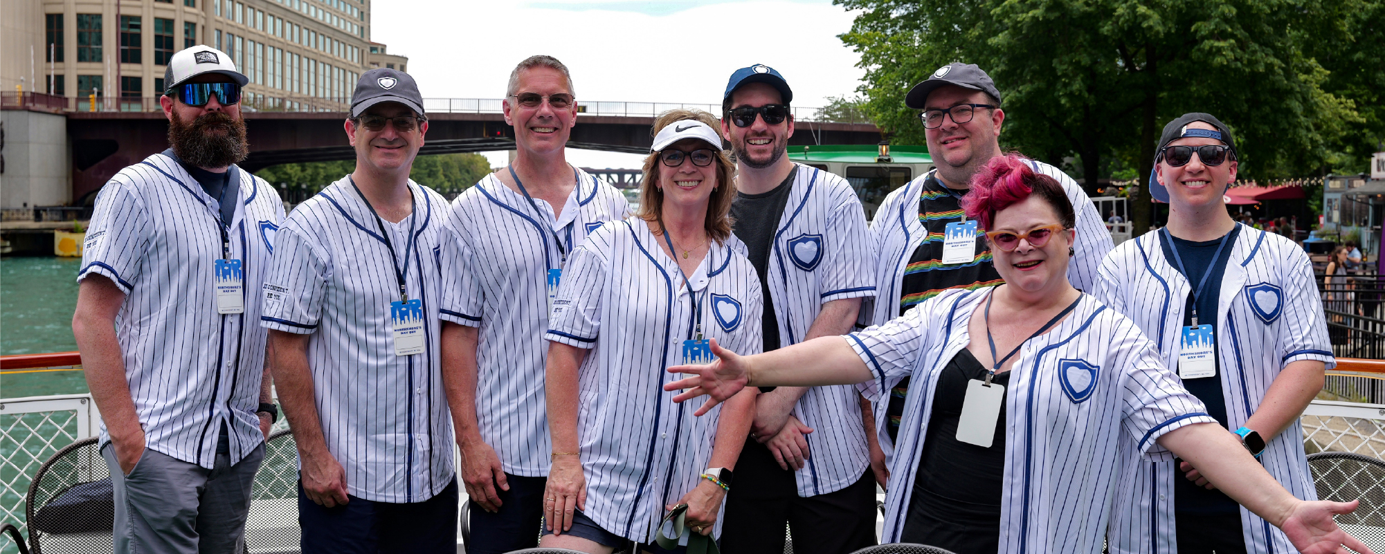 A group of eight adults at NorthShore's Day Out event wearing matching pinstriped baseball jerseys smiles for a photo at a baseball stadium. They stand together near the seating area with the field, scoreboard, and stadium structure visible in the background on a sunny day.