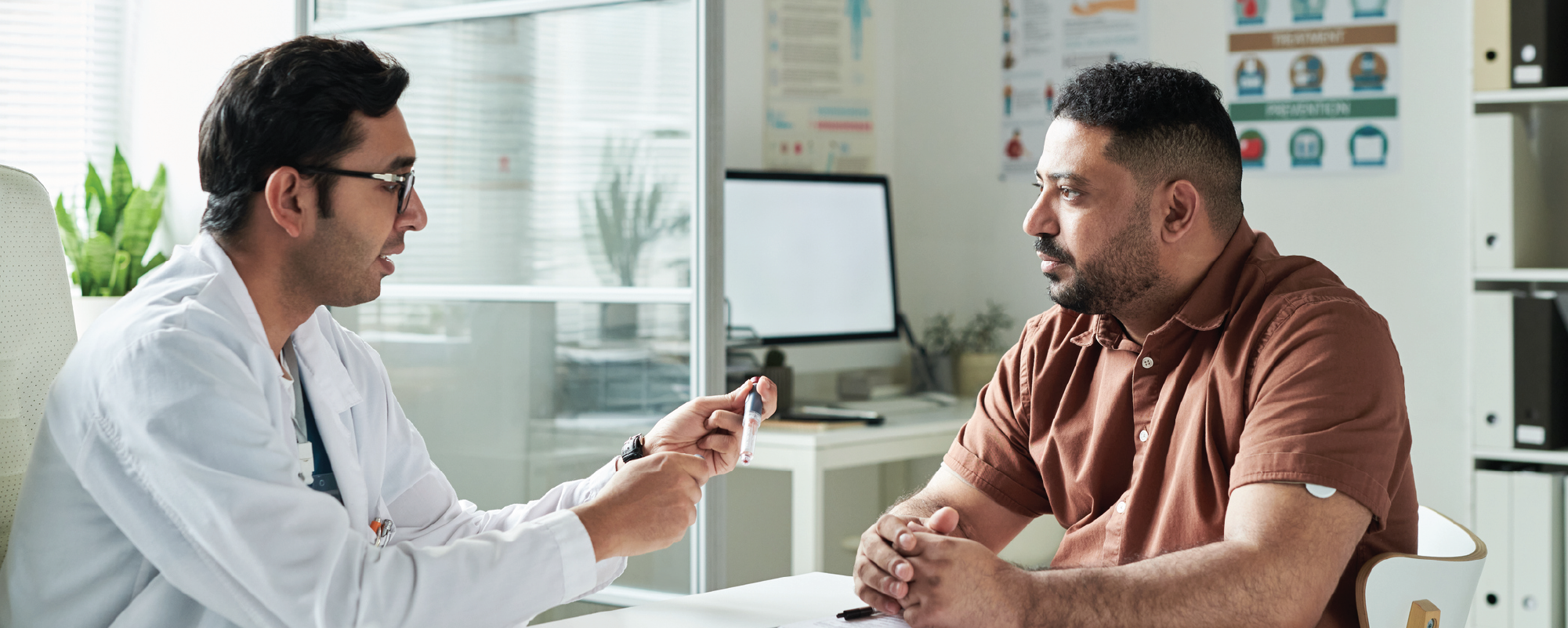 A doctor in a white coat shows a medical device to a patient during a consultation in a modern exam room, as the patient listens attentively with hands clasped on the desk.