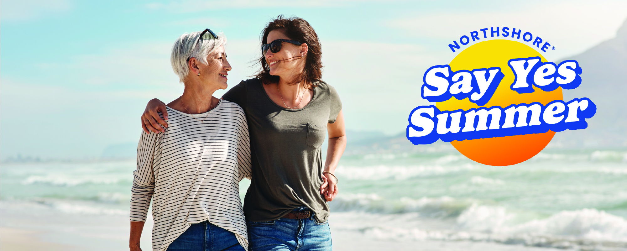 Two women walk arm in arm along a sandy beach, smiling at each other as waves roll in behind them on a sunny day. The upper-right corner features the NorthShore "Say Yes Summer" campaign logo in blue and orange.