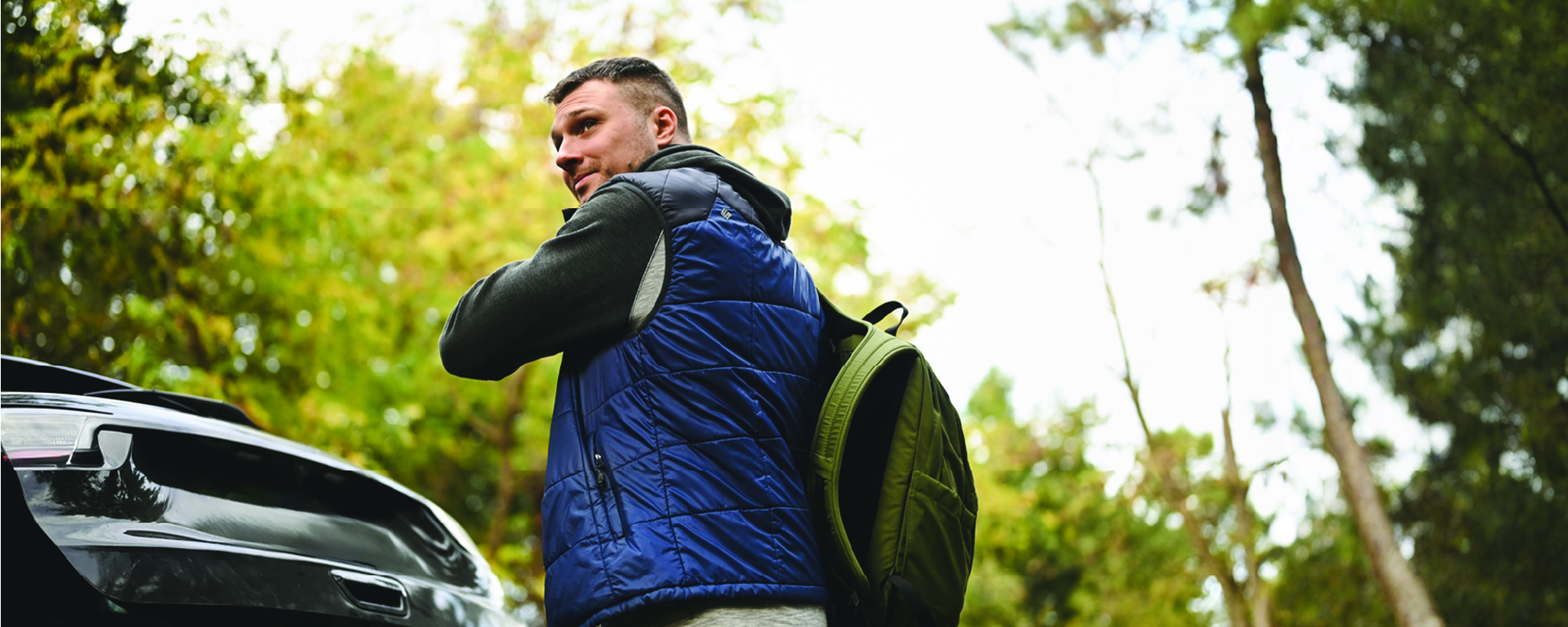 Person wearing a blue quilted vest over a dark hoodie adjusts a backpack while standing beside the open trunk of a car in a wooded area, looking back over their shoulder.