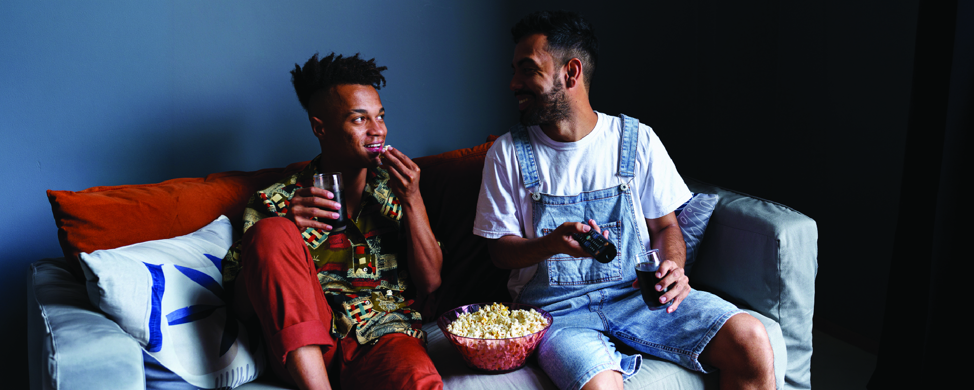Two people sit together on a couch watching something off-camera, sharing popcorn and drinks. One person is holding a game controller while smiling at the other, creating a relaxed, social movie-or-game-night atmosphere. A bowl of popcorn rests between them on the sofa.