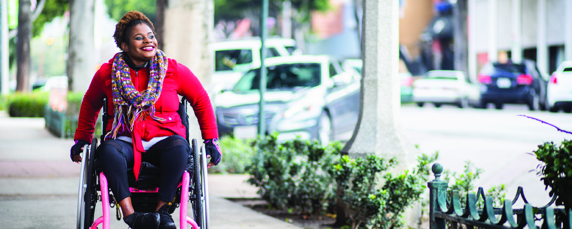 A smiling person using a wheelchair moves along a city sidewalk lined with trees and low shrubs. They are wearing a bright red coat, a colorful patterned scarf, dark pants, and gloves. Parked cars and urban buildings appear blurred in the background.