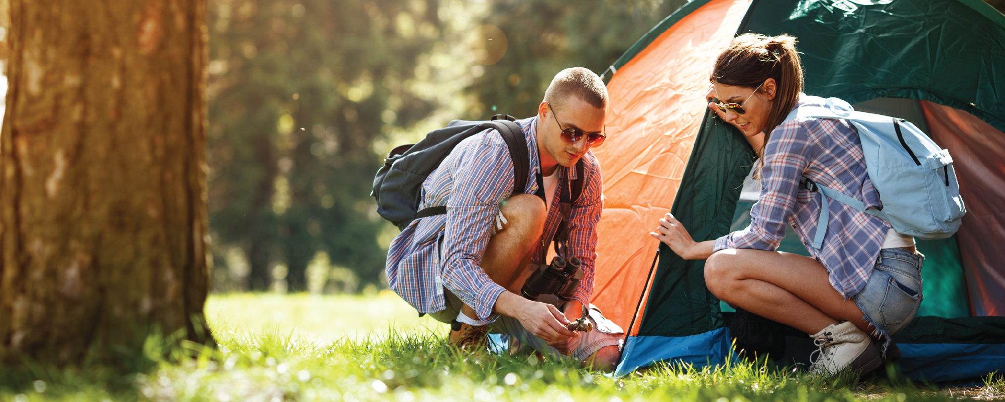 Two campers crouch beside a tent in a sunlit forest, working together to secure it. Both wear casual outdoor clothing and backpacks, with tall trees and soft morning light in the background.