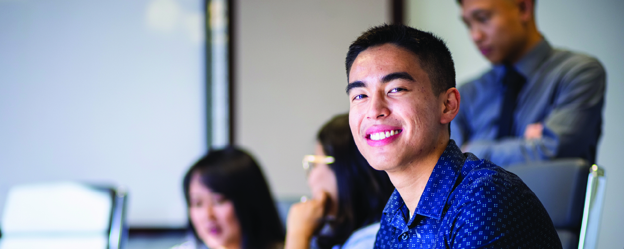 A young man in a blue patterned shirt smiles at the camera while sitting at a desk in a meeting room. Other people in the background are working on laptops and talking, suggesting a collaborative office or classroom setting.