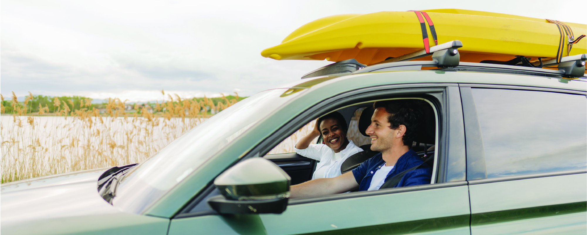 Two people smile from the front seats of a green SUV with two yellow kayaks secured to the roof rack, parked beside a lakeshore with tall reeds in the background.