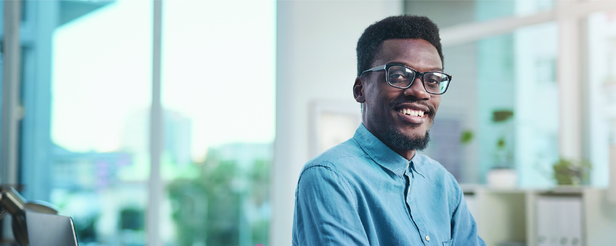 A smiling person wearing glasses and a light blue button-up shirt looks at the camera in a bright office setting. Large windows and blurred shelves in the background create a clean, professional atmosphere.