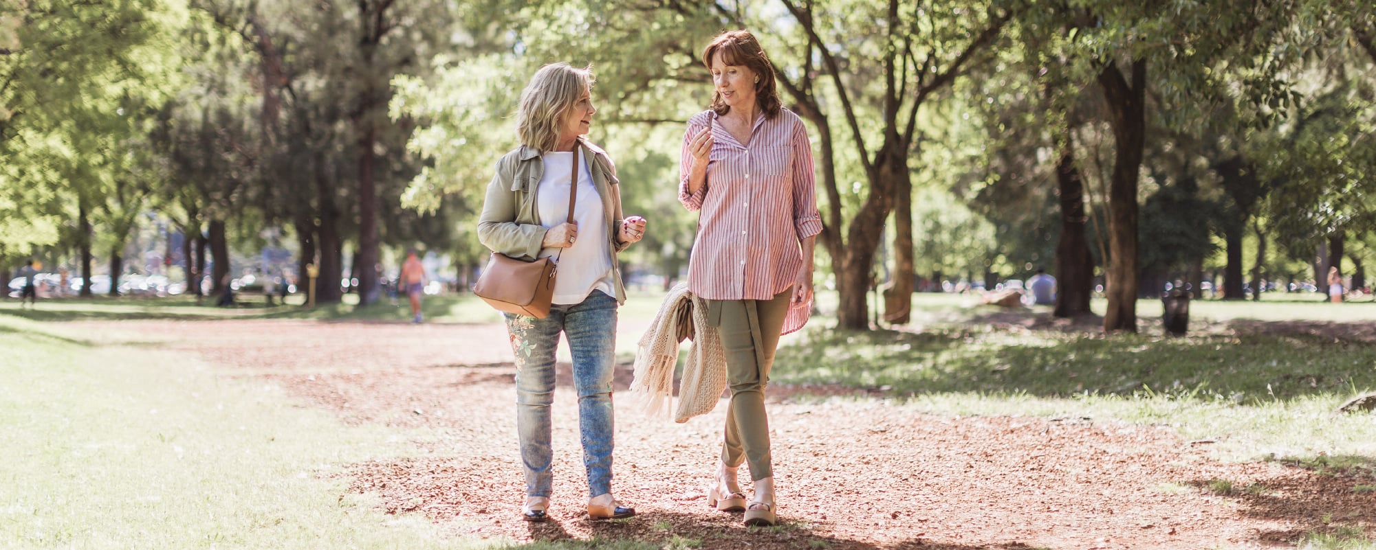Two women walk side by side along a sunlit park path, smiling and talking while carrying light jackets. Tall trees cast dappled shade over the trail, creating a warm, relaxed outdoor setting.