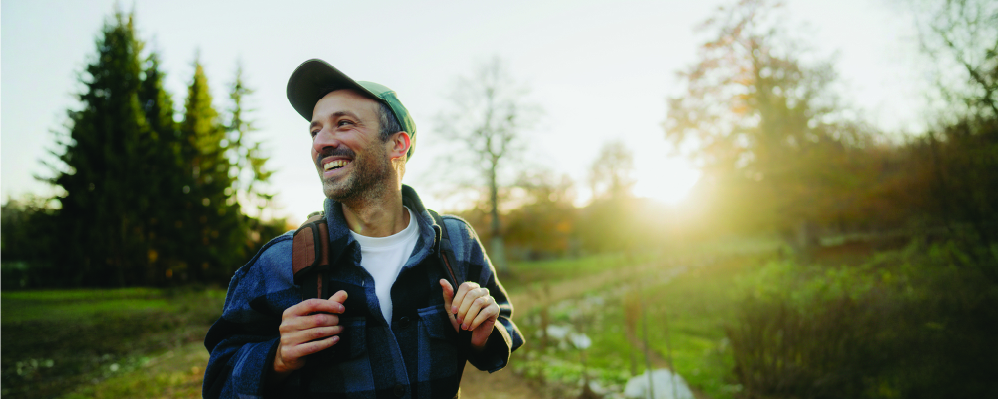 A smiling person wearing a baseball cap and backpack walks along a sunlit nature trail, looking off into the distance. Trees, greenery, and warm golden-hour light create an inviting outdoor setting.