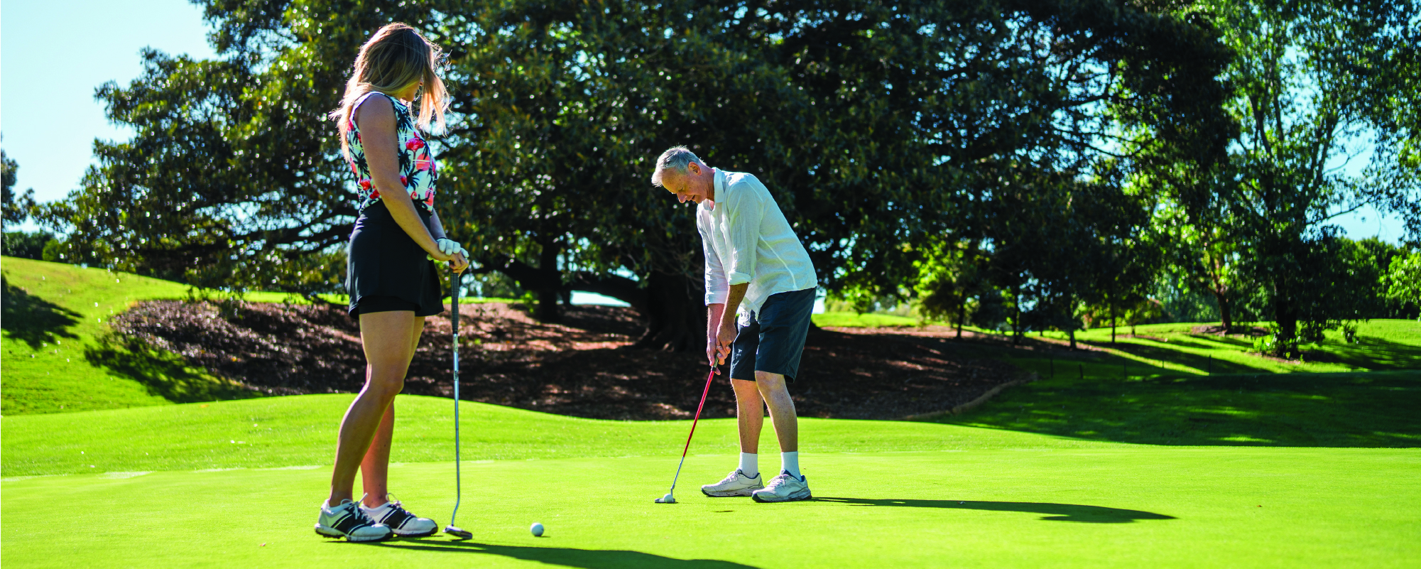 Two people stand on a golf green preparing to putt on a sunny day, with large trees and rolling grass in the background.