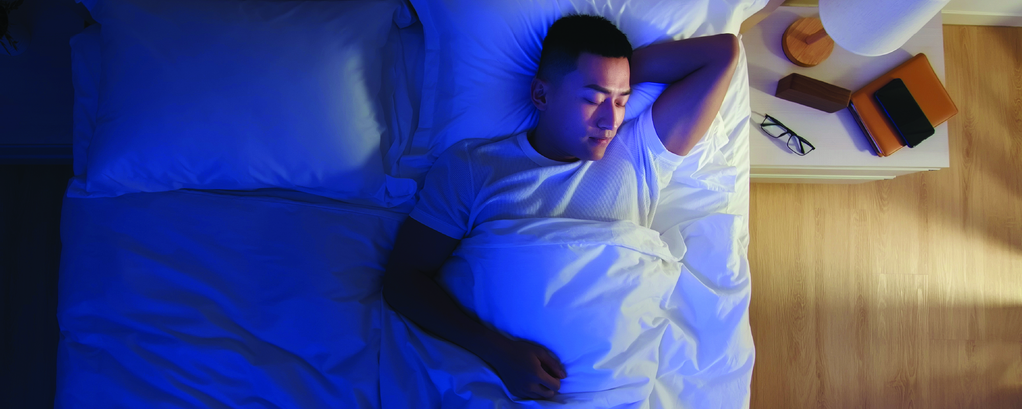 A man lying in bed under white sheets, resting on his back with one arm behind his head, illuminated by soft blue nighttime lighting, with a bedside table and lamp nearby.