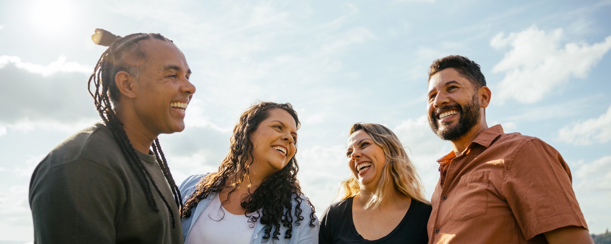 Four adults stand outdoors in a circle, smiling and laughing together under a bright sky. The group appears engaged in a friendly conversation, conveying a sense of connection, support, and community.