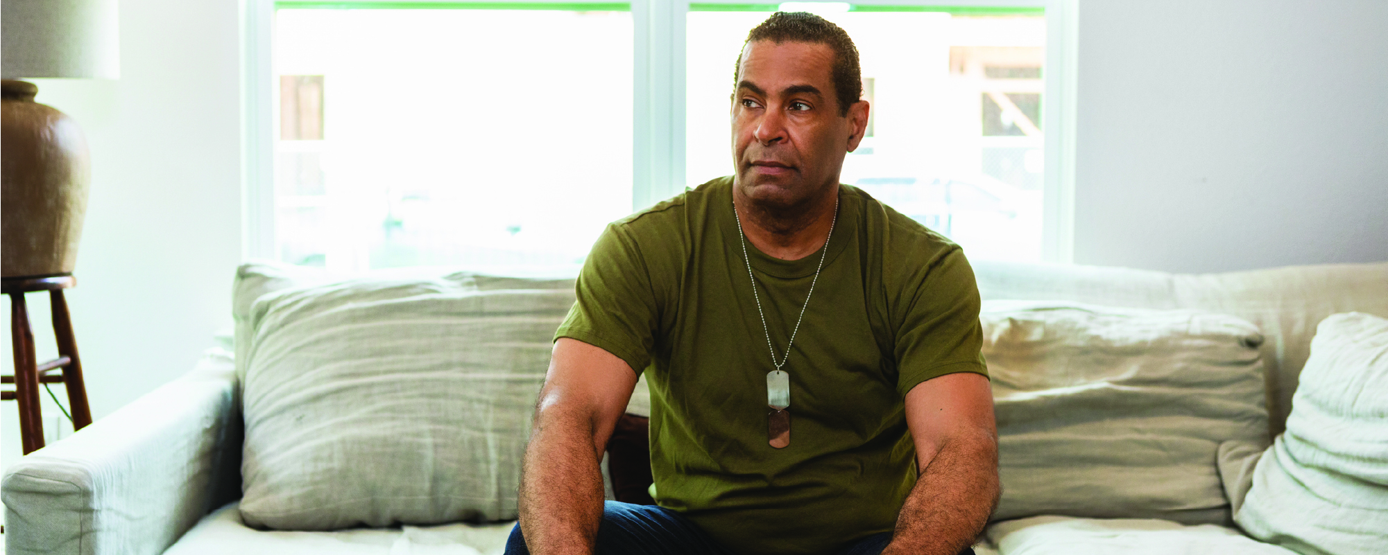 A man wearing a green T-shirt and military-style dog tags sits on a light-colored couch in a bright living room, looking thoughtfully off to the side with his hands resting on his knees.