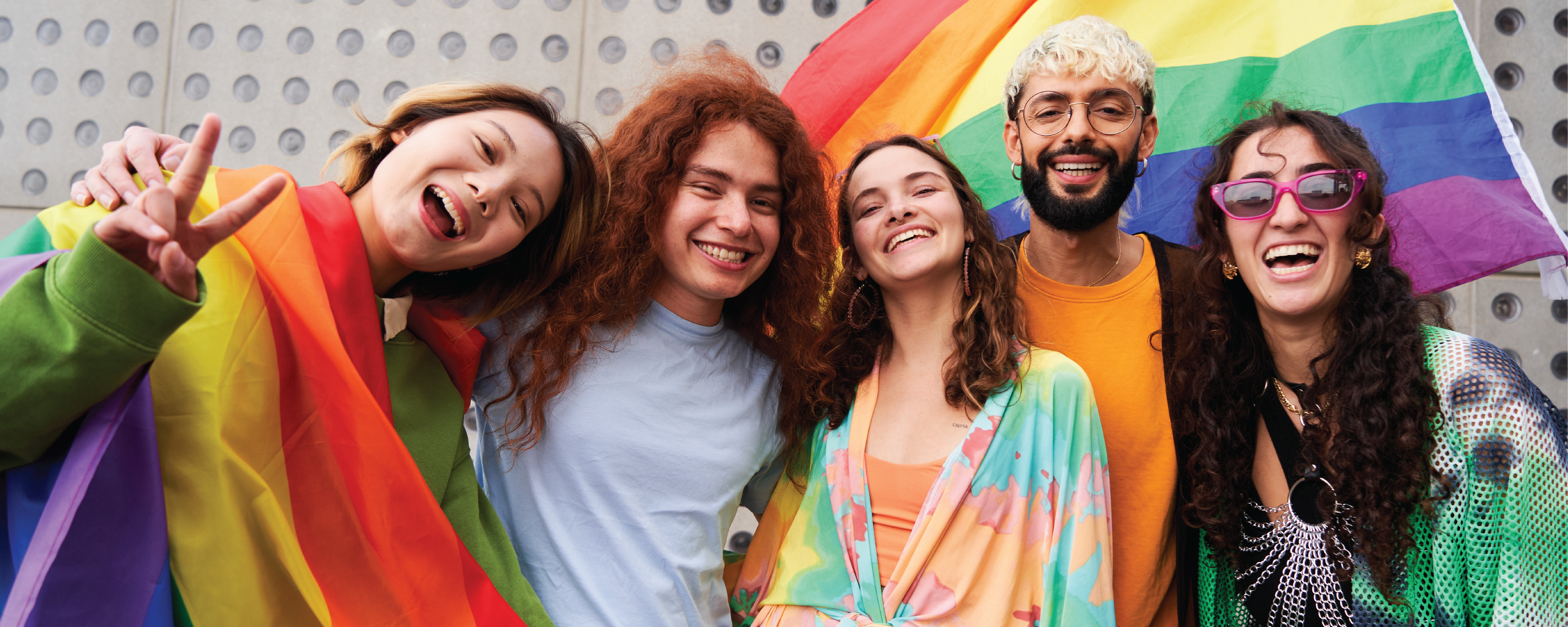 Five smiling people pose together outdoors in front of a large rainbow pride flag. One person is wrapped in a rainbow flag, and the group wears colorful clothing while looking at the camera and celebrating.