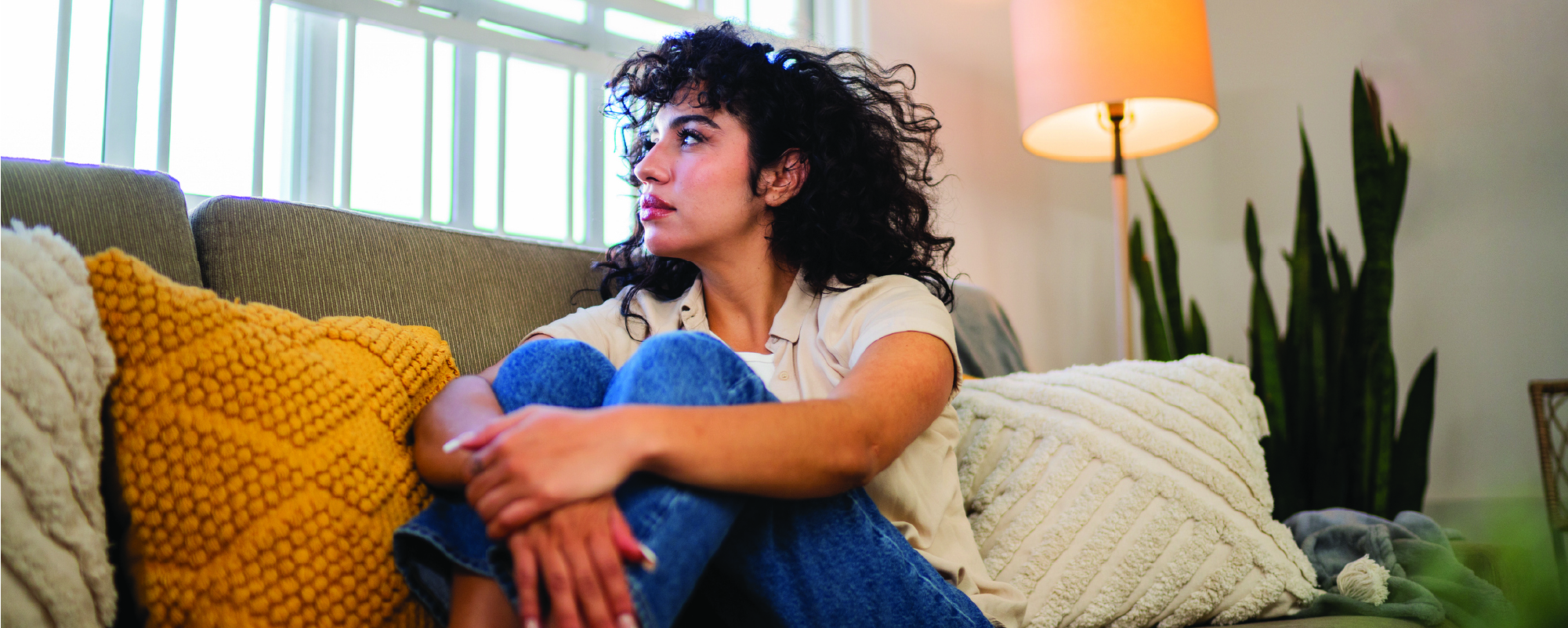 A woman with curly dark hair sits curled up on a couch, looking toward a bright window, with colorful pillows and a lit floor lamp around her.