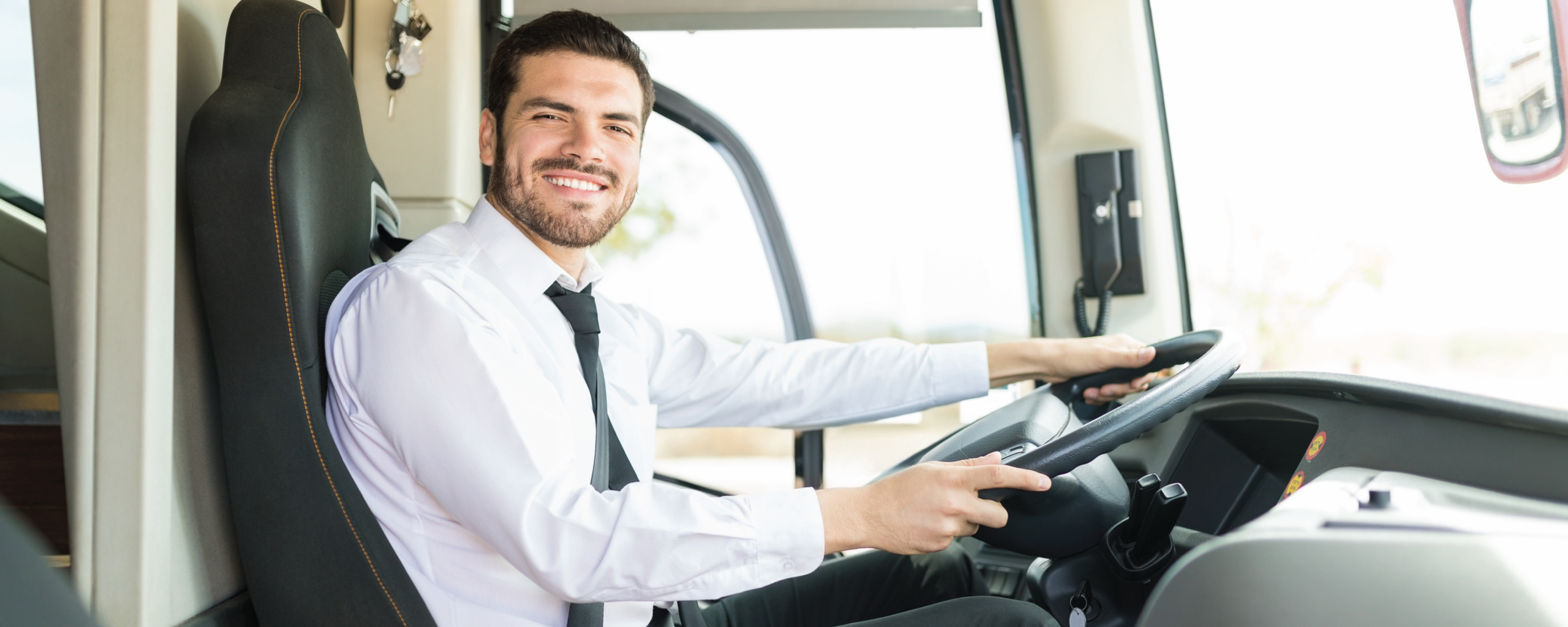 Smiling bus driver wearing a white dress shirt and black tie, seated behind the steering wheel inside a modern bus, looking toward the camera. The driver's seat, dashboard, and large front windows are visible, with bright daylight illuminating the interior.
